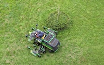 A person operates a green ride-on lawnmower, cutting grass around a small bush on a well-maintained lawn.
