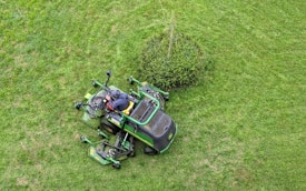 A person operates a green ride-on lawnmower, cutting grass around a small bush on a well-maintained lawn.