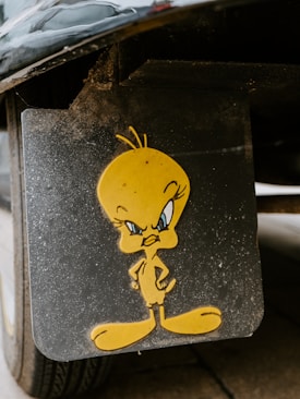 A cartoon character with a yellow body and large feet is depicted on a black surface. The character has a stern expression and stands with its hands on its hips. The surface appears to be part of a vehicle, possibly a mud flap, and shows signs of dirt and wear.