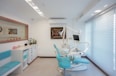 A bright and modern dental clinic room with turquoise dental chairs and equipment. The room is well-lit, featuring a large window with blinds on the right. Cabinets with drawers, decorative plants, and art pieces on the wall enhance its aesthetic appeal.