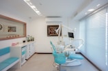 A bright and modern dental clinic room with turquoise dental chairs and equipment. The room is well-lit, featuring a large window with blinds on the right. Cabinets with drawers, decorative plants, and art pieces on the wall enhance its aesthetic appeal.