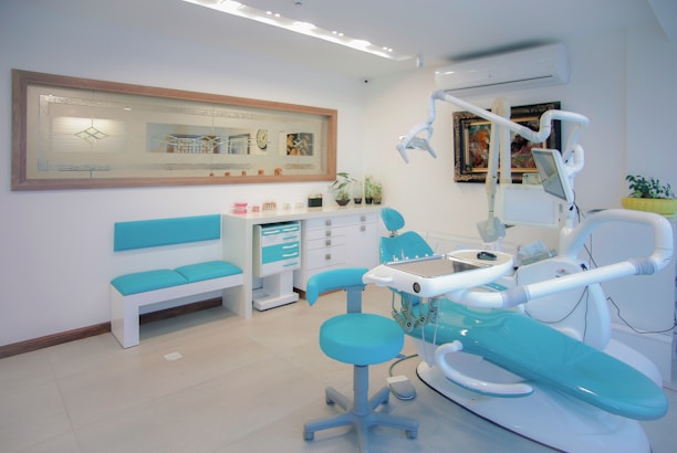 A clean and modern dental office with a dental chair, stools, and various dental equipment in shades of white and teal. The room features a framed artwork, some potted plants, and a bench along one wall. The environment is well-lit with overhead lighting, and there are multiple cabinets and drawers for storage.