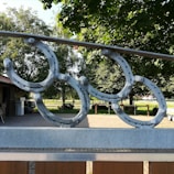 Metal horseshoe decorations are mounted on a fence, situated in an outdoor area with lush green trees and a paved pathway. The background includes a wooden building with a parked vehicle nearby and a bright, sunny landscape.
