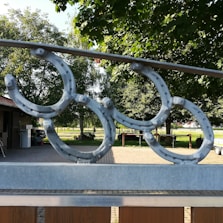 Metal horseshoe decorations are mounted on a fence, situated in an outdoor area with lush green trees and a paved pathway. The background includes a wooden building with a parked vehicle nearby and a bright, sunny landscape.
