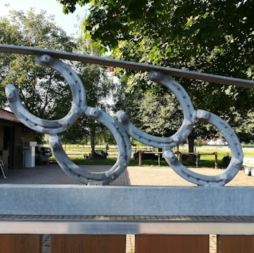 Metal horseshoe decorations are mounted on a fence, situated in an outdoor area with lush green trees and a paved pathway. The background includes a wooden building with a parked vehicle nearby and a bright, sunny landscape.