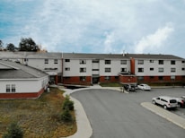 A three-story residential building with a combination of white siding and red brick. Several cars are parked in the parking lot in front of the building. The sky is overcast with patches of blue visible through the clouds. There are a few small evergreen trees on a grassy area adjacent to the building.