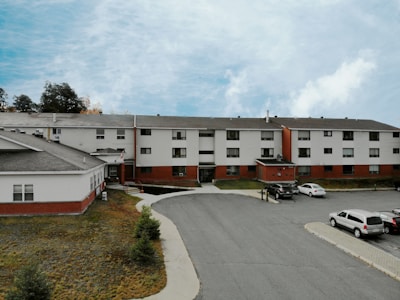 A three-story residential building with a combination of white siding and red brick. Several cars are parked in the parking lot in front of the building. The sky is overcast with patches of blue visible through the clouds. There are a few small evergreen trees on a grassy area adjacent to the building.