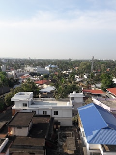 Aerial view of a suburban area featuring numerous buildings with flat and gabled roofs, surrounded by lush greenery and trees. In the foreground, several multi-story houses with white and blue rooftops are visible, while more residential structures can be seen stretching into the distance. The sky is mostly clear with a few clouds.