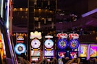 A lively casino scene featuring a row of bright, colorful slot machines with themes like Monopoly, Michael Jackson, and Lord of the Rings. The gaming machines display a variety of flashing lights and digital screens, creating an energetic and exciting atmosphere. Reflections of the lights are visible in glossy surfaces above the machines.