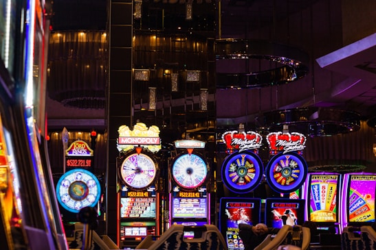 A lively casino scene featuring a row of bright, colorful slot machines with themes like Monopoly, Michael Jackson, and Lord of the Rings. The gaming machines display a variety of flashing lights and digital screens, creating an energetic and exciting atmosphere. Reflections of the lights are visible in glossy surfaces above the machines.