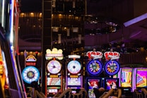A lively casino scene featuring a row of bright, colorful slot machines with themes like Monopoly, Michael Jackson, and Lord of the Rings. The gaming machines display a variety of flashing lights and digital screens, creating an energetic and exciting atmosphere. Reflections of the lights are visible in glossy surfaces above the machines.
