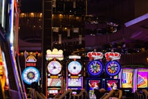 A lively casino scene featuring a row of bright, colorful slot machines with themes like Monopoly, Michael Jackson, and Lord of the Rings. The gaming machines display a variety of flashing lights and digital screens, creating an energetic and exciting atmosphere. Reflections of the lights are visible in glossy surfaces above the machines.