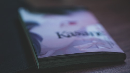A close-up of a slightly closed book or magazine on a wooden surface with an illustration and text on its cover, featuring soft lighting and a shallow depth of field.