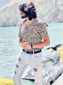 A person with long hair is wearing a leopard print shirt and distressed jeans, standing near a body of water. Behind them is a yellow personal watercraft. The backdrop consists of rocky mountains and clear blue water, suggesting a serene outdoor setting. The person’s posture and the sunny atmosphere contribute to a relaxed vibe.
