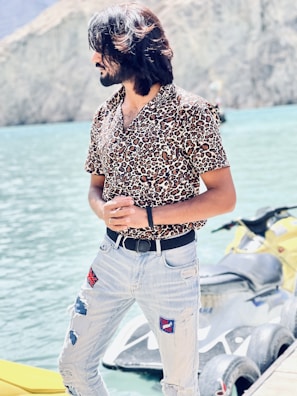 A person with long hair is wearing a leopard print shirt and distressed jeans, standing near a body of water. Behind them is a yellow personal watercraft. The backdrop consists of rocky mountains and clear blue water, suggesting a serene outdoor setting. The person’s posture and the sunny atmosphere contribute to a relaxed vibe.