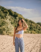 A woman stands on a sandy beach with a forested hillside in the background. She wears a blue patterned crop top and high-waisted jeans. Her long hair is loose, and she holds one hand to her head. The sun casts warm light, highlighting her tattoos.