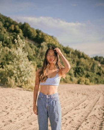 A woman stands on a sandy beach with a forested hillside in the background. She wears a blue patterned crop top and high-waisted jeans. Her long hair is loose, and she holds one hand to her head. The sun casts warm light, highlighting her tattoos.