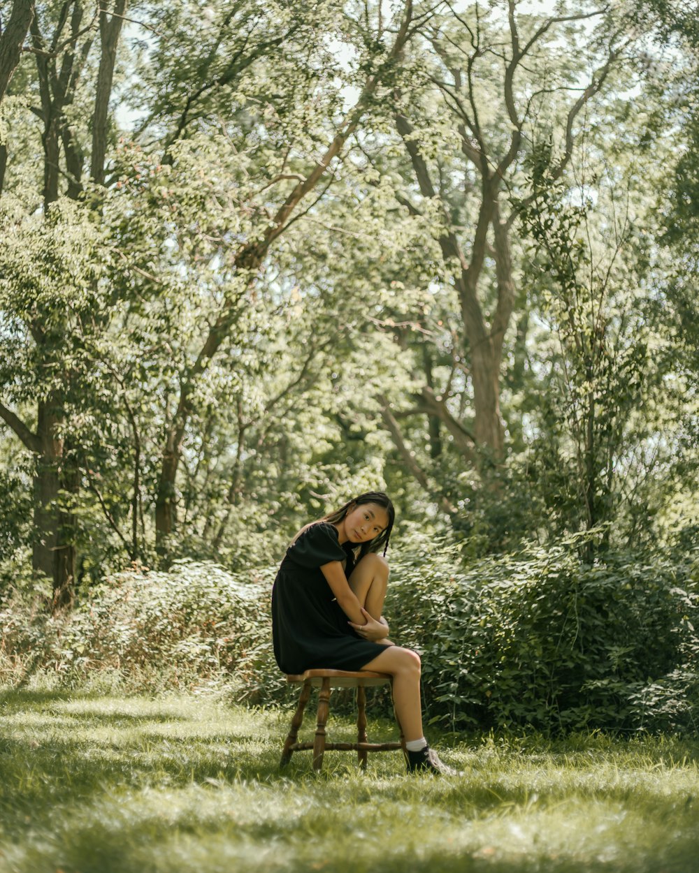 Mujer con camiseta negra sentada en una silla de madera marrón debajo de los árboles verdes durante el día