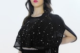 A person is wearing a black embellished garment with intricate embroidery, featuring delicate beadwork and patterns. The attire includes a semi-transparent fabric overlay adorned with decorative designs. The background is plain, highlighting the outfit's details.