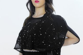 A person is wearing a black embellished garment with intricate embroidery, featuring delicate beadwork and patterns. The attire includes a semi-transparent fabric overlay adorned with decorative designs. The background is plain, highlighting the outfit's details.