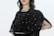 A person is wearing a black embellished garment with intricate embroidery, featuring delicate beadwork and patterns. The attire includes a semi-transparent fabric overlay adorned with decorative designs. The background is plain, highlighting the outfit's details.