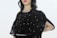 A person is wearing a black embellished garment with intricate embroidery, featuring delicate beadwork and patterns. The attire includes a semi-transparent fabric overlay adorned with decorative designs. The background is plain, highlighting the outfit's details.