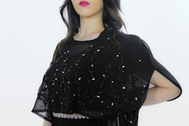 A person is wearing a black embellished garment with intricate embroidery, featuring delicate beadwork and patterns. The attire includes a semi-transparent fabric overlay adorned with decorative designs. The background is plain, highlighting the outfit's details.