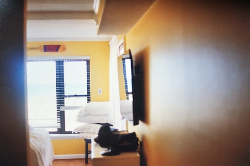 A cozy indoor space with yellow walls, a window with blinds partially open, and folded white towels stacked on a surface. The window light creates a soft and inviting atmosphere.