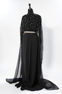 A floor-length black dress displayed on a mannequin. The dress features a flowing, sheer overlayer adorned with subtle embellishments, and a prominent decorative belt at the waist. The design is elegant and sophisticated, suitable for formal occasions.
