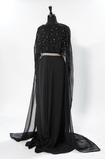 A floor-length black dress displayed on a mannequin. The dress features a flowing, sheer overlayer adorned with subtle embellishments, and a prominent decorative belt at the waist. The design is elegant and sophisticated, suitable for formal occasions.