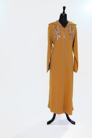 A mustard-colored garment with intricate floral embroidery on the upper chest, displayed on a black mannequin. The garment features long sleeves and a necklace accessory.