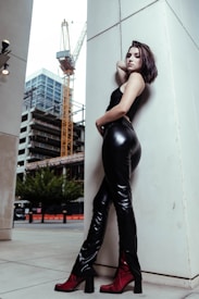 A person in black leather attire poses confidently against a large white column. In the background, there's a construction site with a crane and a partially constructed building, suggesting an urban environment. The person wears striking red shoes, adding a vibrant contrast to the otherwise muted color palette.