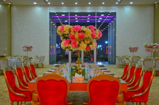 A lavishly decorated banquet hall featuring a long dining table set for a formal event. The table is adorned with a grand floral centerpiece consisting of pink and white flowers. Surrounding the table are ornate chairs with deep red upholstery. The room is illuminated with bright overhead lighting, and several round tables with similar decor are visible in the background.