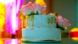 A two-tiered white cake adorned with pink roses and gold drips sits elegantly on a reflective surface. The initials 'D&Z' are inscribed on the side, adding a personalized touch. The background is vibrant and colorful, with shades of yellow and pink creating a warm ambiance.
