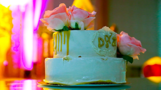A two-tiered white cake adorned with pink roses and gold drips sits elegantly on a reflective surface. The initials 'D&Z' are inscribed on the side, adding a personalized touch. The background is vibrant and colorful, with shades of yellow and pink creating a warm ambiance.
