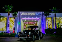 An elegant building with illuminated palm trees stands as the backdrop for a vintage black car parked in front. The entrance is adorned with bright lights and signage that reads 'Salle des f&ecirc;tes' with decorative stars. The scene is vibrant with a variety of colors highlighting the building's modern architecture.