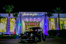 An elegant building with illuminated palm trees stands as the backdrop for a vintage black car parked in front. The entrance is adorned with bright lights and signage that reads 'Salle des fêtes' with decorative stars. The scene is vibrant with a variety of colors highlighting the building's modern architecture.