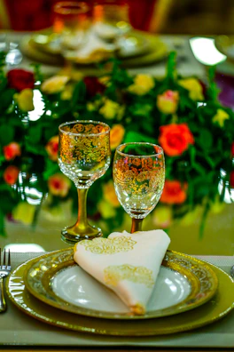 Elegant table setting with two gold-rimmed wine glasses and fancy dinner plates, accompanied by a white napkin with gold embroidery. In the background, vibrant flowers in shades of red, yellow, and green create a lush centerpiece.