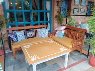 An outdoor seating area featuring two wooden benches with beige cushions and a variety of colorful patterned throw pillows. A rectangular table with decorative white patterns sits in front of the benches. The scene is framed by a blue window with plants on either side, and wall decor including framed pictures and a lantern.