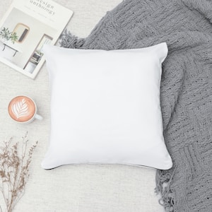 A white pillow lies on a textured gray blanket next to a cup of latte art coffee and a magazine titled 'Design Anthology'. Small dried plants are also visible, adding a touch of nature to the cozy scene.