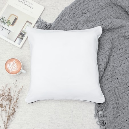A white pillow lies on a textured gray blanket next to a cup of latte art coffee and a magazine titled 'Design Anthology'. Small dried plants are also visible, adding a touch of nature to the cozy scene.