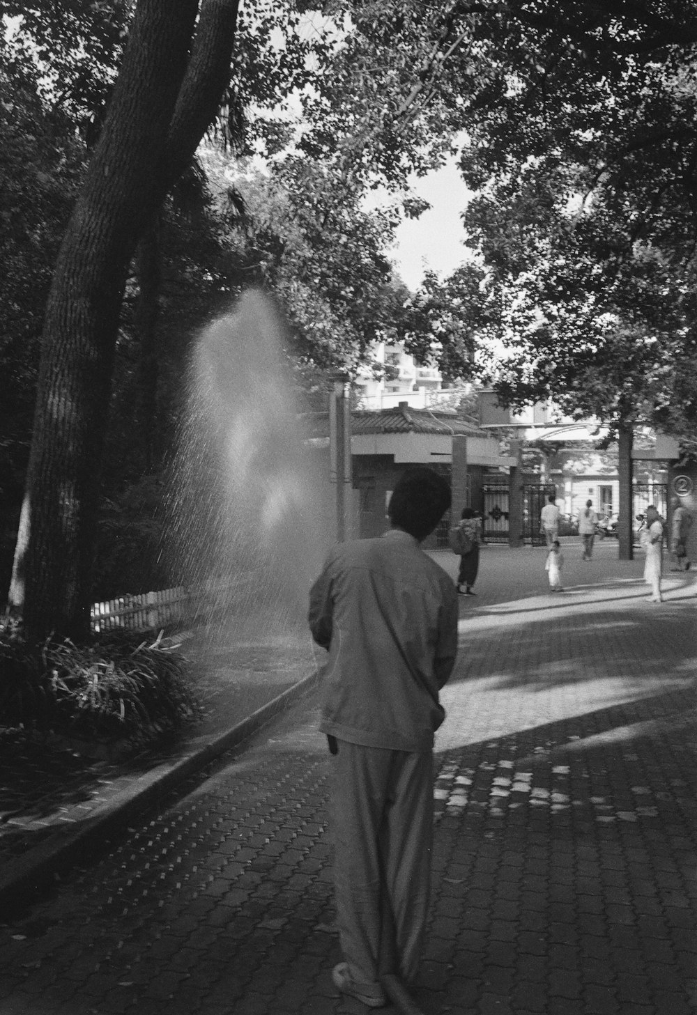 man in black coat standing on sidewalk near water fountain in grayscale photography