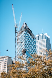 A modern skyscraper under construction with a large crane alongside it. The building features a glass facade with geometric patterns and scaffolding at the top. In the foreground, there are trees with golden leaves, and another completed skyscraper is visible on the right.