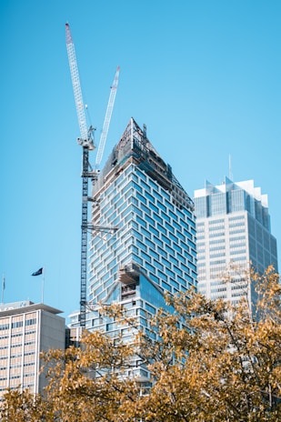 A modern skyscraper under construction with a large crane alongside it. The building features a glass facade with geometric patterns and scaffolding at the top. In the foreground, there are trees with golden leaves, and another completed skyscraper is visible on the right.