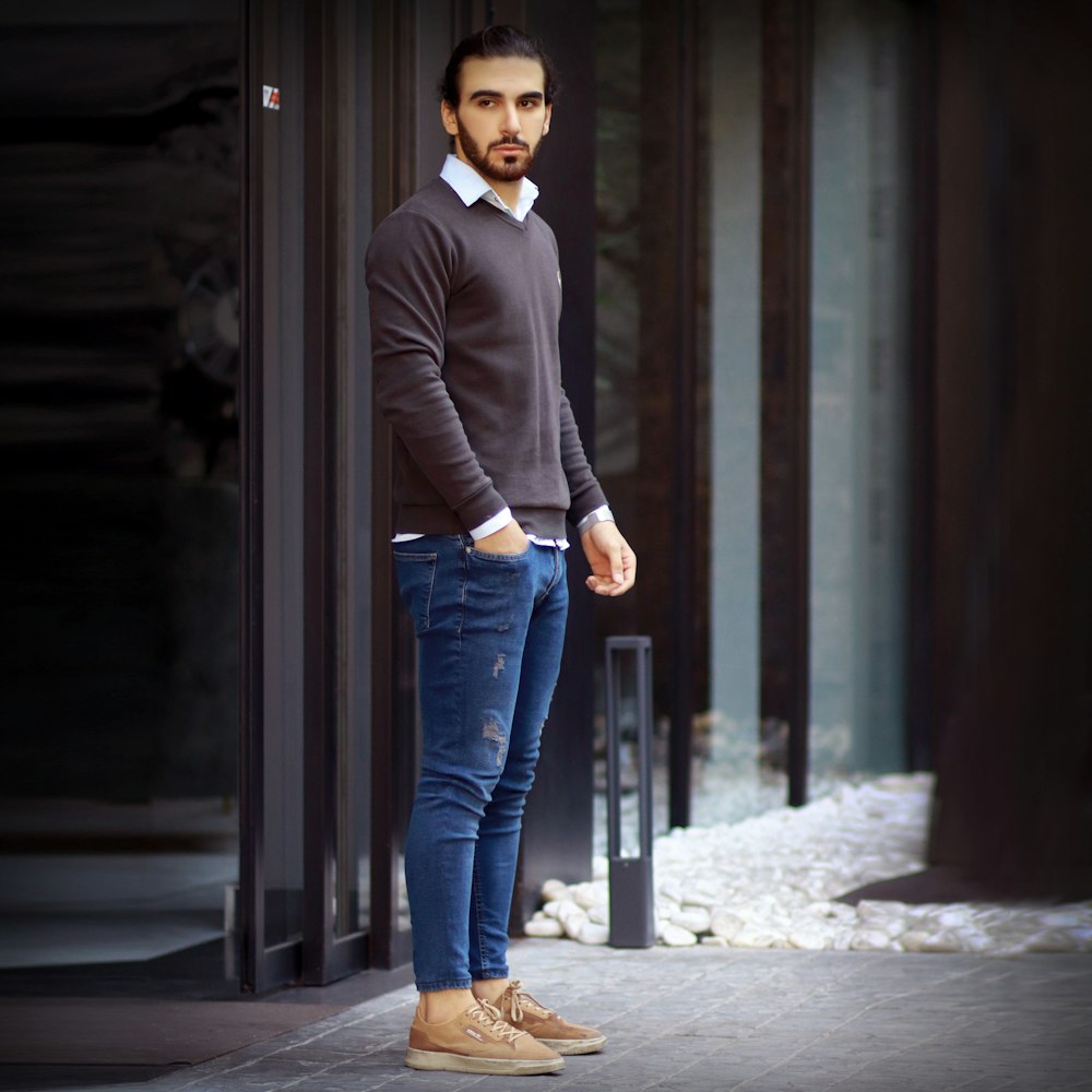 man in black blazer and blue denim jeans standing beside brown wooden door during daytime