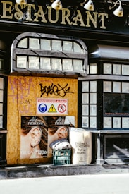 A boarded-up section of a building features large lettering indicating a restaurant. The boarded area displays graffiti and three safety signs depicting head protection, a warning, and restricted access. Two identical posters show an ear with piercings. A plastic crate with a folded cloth and a sack with printed text suggesting cleanliness for Paris are placed nearby.