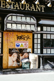 A boarded-up section of a building features large lettering indicating a restaurant. The boarded area displays graffiti and three safety signs depicting head protection, a warning, and restricted access. Two identical posters show an ear with piercings. A plastic crate with a folded cloth and a sack with printed text suggesting cleanliness for Paris are placed nearby.