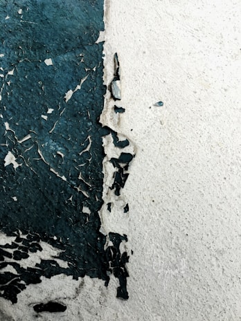 Peeling paint on a wall reveals a rough texture beneath. The paint is dark green in some areas, creating an abstract and weathered appearance against the lighter wall surface.