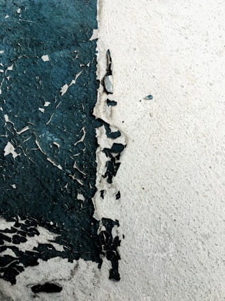 Peeling paint on a wall reveals a rough texture beneath. The paint is dark green in some areas, creating an abstract and weathered appearance against the lighter wall surface.