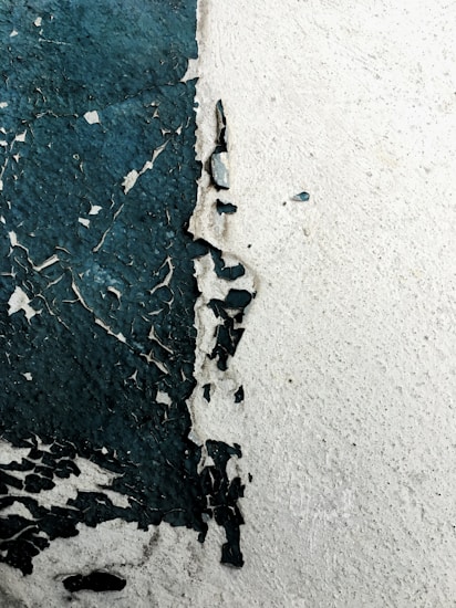 Peeling paint on a wall reveals a rough texture beneath. The paint is dark green in some areas, creating an abstract and weathered appearance against the lighter wall surface.